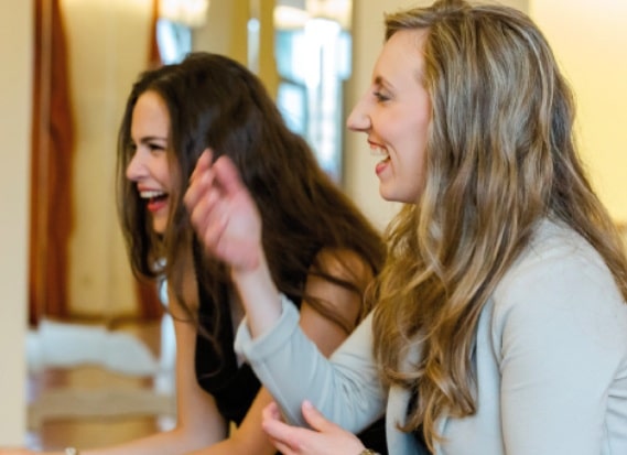 Zwei junge Frauen mit langen Haaren sitzen nebeneinander, lächeln und lachen zusammen in einem warm beleuchteten Innenraum.